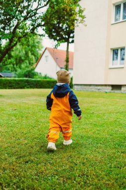 Parlak turuncu yağmurluk giymiş neşeli bir çocuk, uzun ağaçlarla çevrili yemyeşil bir çimenliği ve açık havada saf neşe ve merakı somutlaştıran büyüleyici bir evi zevkle keşfeder.