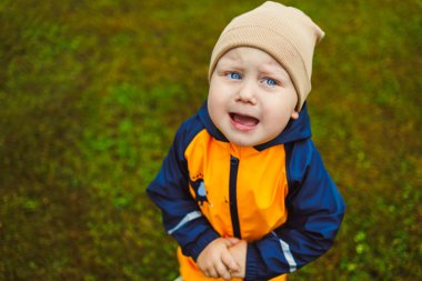Parlak yağmurluk ve rahat şapkalı neşeli bir çocuk yeşil çimlerin üzerinde duruyor, güzel bir açık hava ortamında duygularını neşeyle ifade ediyor, keşif ve oyun için mükemmel.
