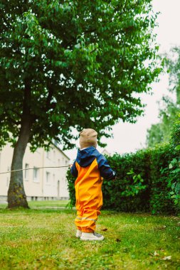 Canlı turuncu ve mavi yağmurluk giymiş genç bir çocuk, bereketli bir günde yemyeşil bir bahçeyi neşeyle keşfediyor, yemyeşil yapraklar ve güzel bir ağaçla çevrili.