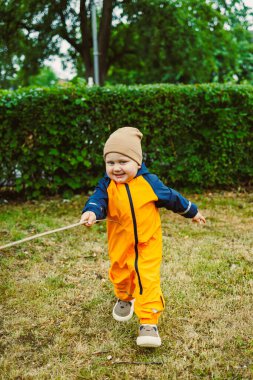Parlak turuncu yağmurluk ve bej şapkalı neşeli bir çocuk yeşil çimenlerde koşar, kaygısız çocukluk, macera ve açık havada canlı bir sahnede oynar.