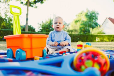 Güneşli bir parkta parlak oyuncaklarla çevrili bir battaniyenin üzerinde oturan sevimli bir çocuk, dışarıda oyun oynarken neşe ve masumiyetin somutlaştırılmış hali.