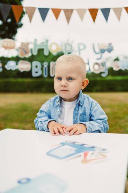 Canlı bir açık hava doğum günü partisinde, kot ceketli neşeli sarışın bir çocuk canlı dekorasyonların tadını çıkarıyor, eğlence ve heyecan dolu neşeli bir ortam yaratıyor.