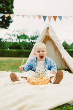 Neşeli bir bebek açık havada yumuşak bir battaniyeye oturur, neşeli bir ifadeyle atıştırmalıkların tadını çıkarır, renkli kiraz kuşu ve rahat bir çadır ile çevrili, neşeli havayı güçlendirir.