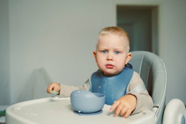Parlak mavi gözlü neşeli bir çocuk yemek masasında yüksek bir sandalyede oturuyor, sevgi dolu bir aile ortamında renkli bir kâse ve kaşıkla neşeyle oynuyor.