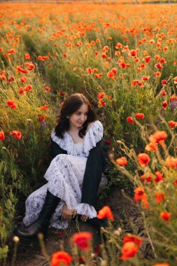 Dalgalı beyaz elbiseli bir kadının turuncu gelinciklerle dolu nefes kesici bir tarlada oturduğu sakin bir sahne gün batımında doğanın güzelliğinin özünü yakalıyor..