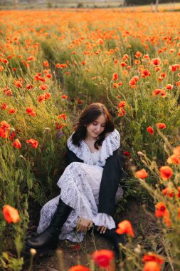 Parlak turuncu gelincikler arasında zarif bir şekilde oturan beyaz elbiseli bir kadının huzur ve doğal güzellik hissi yaydığı sakin bir sahne.