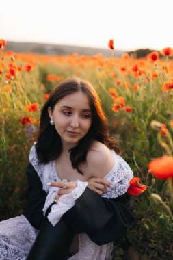 Genç bir kadın haşhaş çiçeklerinden oluşan canlı bir denizin ortasında zarif bir şekilde oturuyor. Güneş etrafındaki çiçek açan tarlanın üzerinde güzel bir şekilde batarken derin bir yansımanın tadını çıkarıyor.