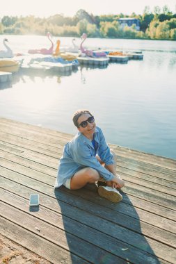 Neşeli genç bir kadın nehir kenarında dinleniyor, sıcak güneş ışığının tadını çıkarırken renkli yüzgeçli kayıklar yakınlarda yüzüyor, hareketli sahneyi güzelleştiriyor.