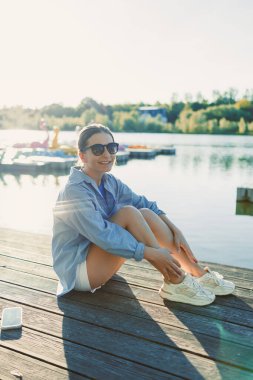 Şık güneş gözlüklü neşeli bir kadın sakin bir gölün kenarındaki ahşap bir rıhtımda güneşleniyor. Huzur, mutluluk ve rahatlık dolu mükemmel bir günün tadını çıkarıyor.