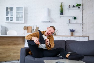 Neşeli genç bir adam, şık, modern bir oturma odasında rahat, keyifli ve eğlenceli bir atmosfer sunan rahat bir kanepede otururken neşeli bir şekilde kollarını uzatır.