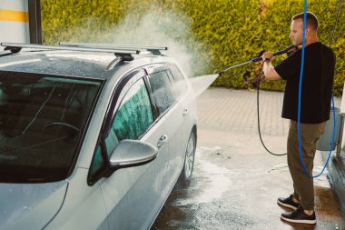 Bir adam arka planda yemyeşil yeşilliklerle çevrili bir oto yıkamada yüksek basınçlı su spreyiyle gümüş bir arabayı aktif olarak yıkıyor.