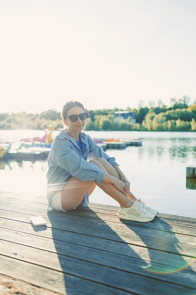 Genç bir kadın rıhtımda dinleniyor, sakin bir gölde sıcak güneş ışığının tadını çıkarıyor, güzel bir yaz gününde gevşemek ve hayatın tadını çıkarmak için mükemmel bir ortam yaratıyor.