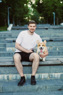 Güzel ve güneşli bir günde, neşeli genç bir adam parkın merdivenlerinde sevimli ve neşeli Corgisiyle otururken, saf neşe ve dostluk sergiliyor.