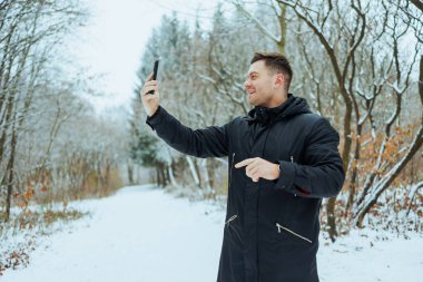 Neşeli bir adam, uzun, karla kaplı ağaçlarla çevrili, karlı bir ormanda kışın getirdiği huzurlu sükunetin tadını çıkaran neşeli bir selfie çeker.