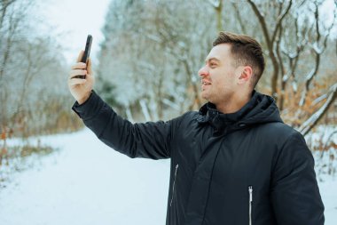 Neşeli genç bir adam güzel, karlı bir ormanda neşeli bir şekilde dikiliyor, akıllı telefonuyla heyecan verici bir selfie çekerken kışın büyüleyici güzelliğini gösteriyor.