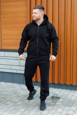 Siyah kapüşonlu ve eşofmanlı, kendinden emin genç bir adam modern ahşap arka planda rahatlamış ama şık bir görünüm sergiliyor.