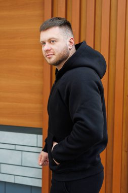 Şık, genç bir adam şık, siyah bir kapüşonluyla poz veriyor. Modern, güzel, ahşap bir arka plana bakıyor. Görsel estetiği geliştiriyor.