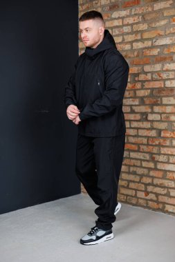 Modern bir endüstriyel ortamda, erkek model şık bir siyah atletizm kıyafeti giyer, izleyicileri büyüleyen ve kalıcı bir etki bırakan çarpıcı bir görünüm yaratır.