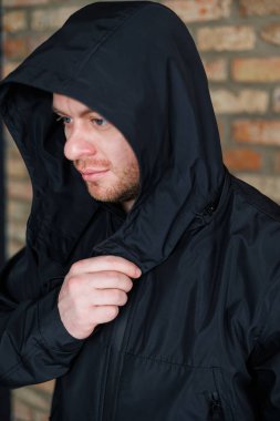 Yakından bakınca, şık kapüşonlu, parlak siyah su geçirmez ceketli bir adamın, şık görünümünü, dokulu tuğla bir duvara karşı geliştirdiğini, kentsel bir hava sergilediğini görüyoruz.