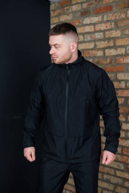 Şık bir genç adam, parlak siyah bir ceket ve uyumlu pantolon giyen, cesur bir moda ve karizma yayarak, dokulu tuğladan bir arka plana karşı güven içinde poz verir.