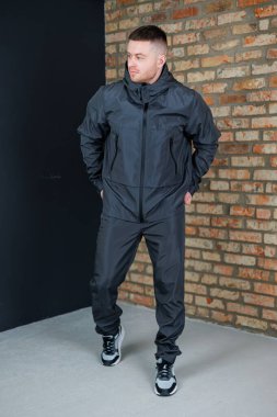 Parlak siyah atletizm kıyafeti giymiş bir adam dokulu tuğla bir duvarın yanında duruyor ve aktif yaşam tarzları için en son moda akımlarını sergiliyor.