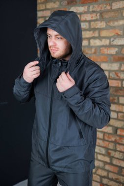 Modern, koyu renk kapüşonlu şık bir adam kırsal tuğla bir duvara karşı düşünceli bir duruş sergiliyor, fevkalade ve eşsiz şehir moda anlayışını oldukça iyi sergiliyor.