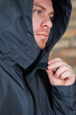 Bu yakın planda, düşünceli bir şekilde koyu renk ceketinin kaputunu düzelten bir adam görüyoruz. Düşünceli bir yüz ifadesi sergiliyor.