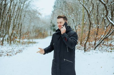 Siyah kışlık ceketli bir adam akıllı telefonuyla konuşuyor, sakin bir kış manzarasında karlı bir yolda çıplak ağaçlarla yürüyor, güzel bir soğuk mevsimi sahnesi yaratıyor.