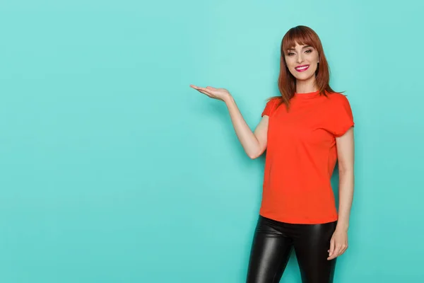Smiling young woman in orange shirt is holding hand raised, showing something and looking at camera. Three quarter length studio shot on turquoise background.