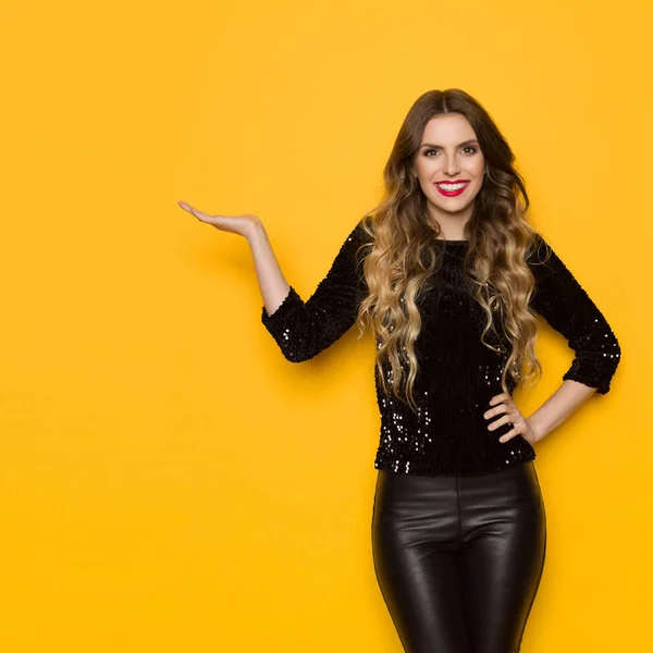 Beautiful young woman in black sequin sweater and leather pants is standing with hand raised, showing something and smiling. Three quarter length studio shot on yellow background.