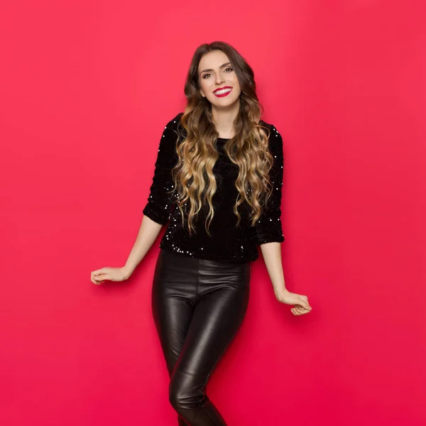 Beautiful young woman in black sequin sweater and leather pants is smiling and looking at camera. Three quarter length studio shot on red background.