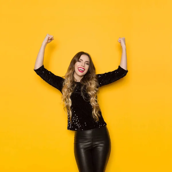 Beautiful young woman in black sequin sweater and leather pants is shouting with arms raised. Three quarter length studio shot on yellow background.