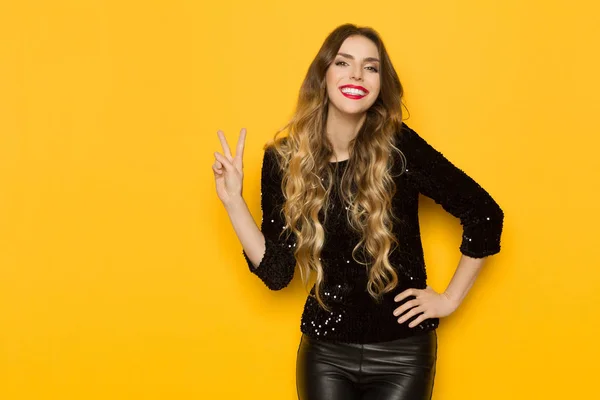 Beautiful young woman in black sequin top and leather trousers is showing peace hand sign and smiling. Three quarter length studio shot on yellow background.