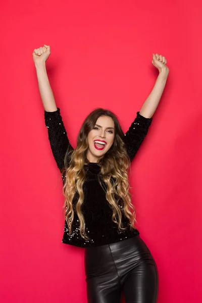 Beautiful young woman in black sequin sweater and leather pants is raising arms and laughing. Three quarter length studio shot on red background.