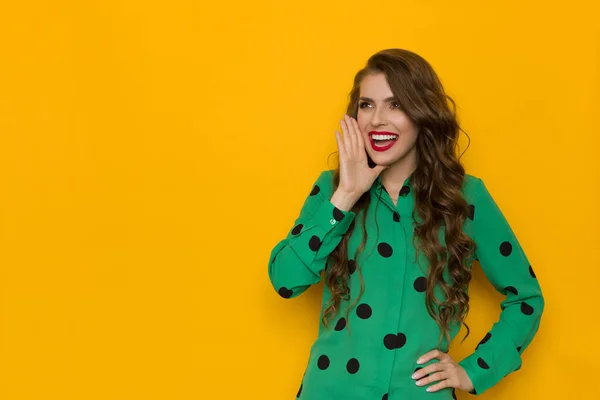 Excited young woman in green shirt in black dots is holding hand on chin, looking away and shouting. Waist up studio shot on yellow background.