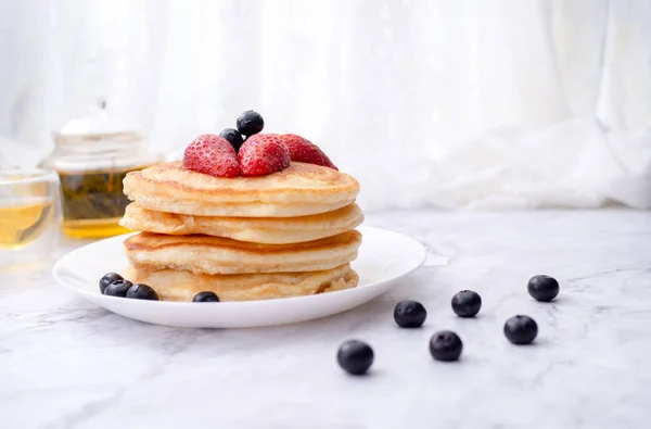 Stack di frittelle con guarnizione, fragola e mirtillo.Posizionato in un piatto bianco su un tavolo di marmo e copiare spazio.Mangiare con il tè nel bicchiere . — Foto Stock