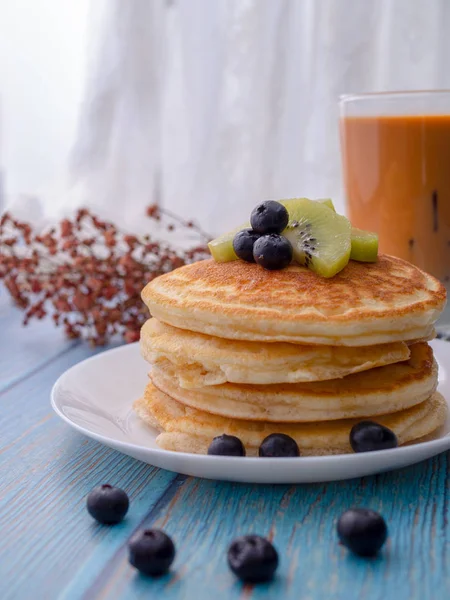 Stapel pannenkoeken met topping, kiwi en Blueberry. geplaatst in een witte plaat op een blauwe houten tafel. Eten met melk thee in het glas. — Stockfoto