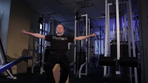 Un homme de l'âge effectue une gymnastique articulaire pour les doigts dans la gymnase. Serrer et déserrer votre poing, bras étendus à gauche et à droite. Copier l'espace — Video