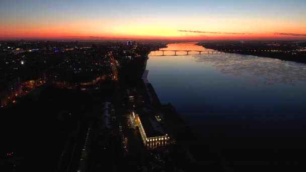 Vista aérea oscura de la ciudad nocturna en un amplio río con un puente al atardecer. La puesta de sol se refleja en el agua. — Vídeos de Stock