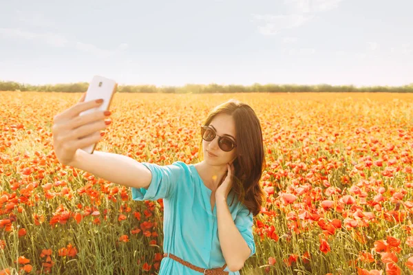 日当たりの良い夏の日の赤いケシ草原の背景にスマート フォンで Selfie を取って美しい若い女性 — ストック写真