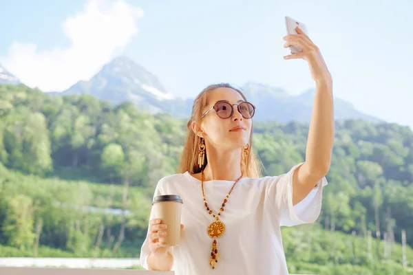夏の山の背景にバルコニーでスマート フォンで Selfie を取ってコーヒーのカップを持つ若い女性を笑顔 — ストック写真