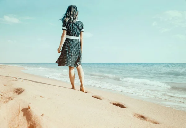 背面のそばの足跡を残して砂のビーチの上を歩くドレスを着た裸足の若い女性 夏の休暇 — ストック写真