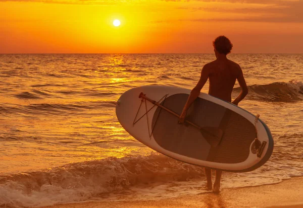 Young man with SUP board going to the sea at sunset, beach vacations.