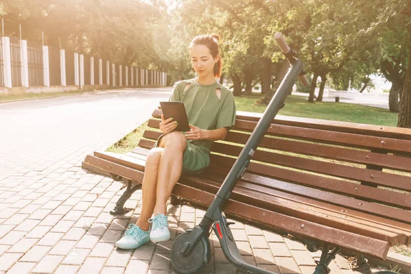 Chica descansando en el banco con la tableta digital y un e-scooter en pa —  Fotos de Stock