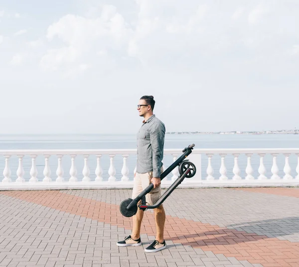 Man holding folding electric scooter on city embankment. — Stock Photo, Image