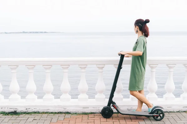 Gadis berdiri dengan skuter listrik dan melihat laut — Stok Foto