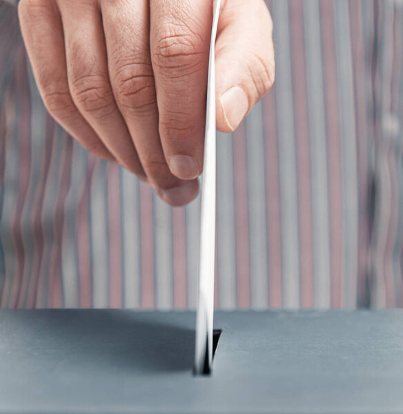 Мужская рука кладет бюллетень в урну для голосования крупным планом. Выборы
.