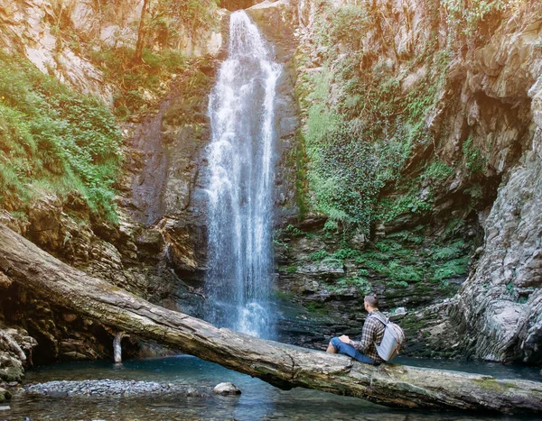 Explorer young man with backpack resting on fallen tree trunk and enjoying view of waterfall in summer.