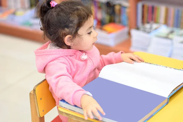 어린 소녀가 책 앞에 앉아 있다. 식탁 과 책을 읽는 곳 근처 의자에 앉아 있는 어린 아이를 귀엽게 대한다. — 스톡 사진