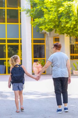 Büyükanne ve öğrenci çocuk okula giderken el ele tutuşuyor. Okul çantalı küçük kız ya da büyükannesiyle okula yürüyen çantalı kız. Yaşlı ebeveyn ve kız, büyükanne ve torun, okula dönüyorlar..